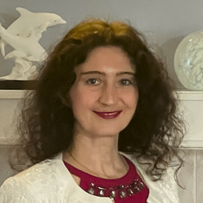 A woman with curly brown hair and red lipstick smiles at the camera. Wearing a white cardigan over a red top with a decorative necklace, she stands before a shelf with a white dolphin sculpture and glass orb, reflecting on teacher retention.